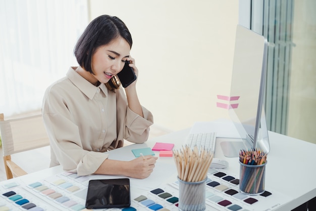 Concepteur de belle femme asiatique, parler au téléphone avec le client et prendre note des détails du travail