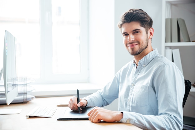 Concepteur de jeune homme heureux assis et utilisant une tablette graphique sur le lieu de travail