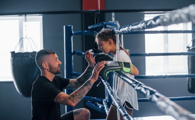 Conception de l'aide. Le jeune entraîneur de boxe tatoué enseigne la petite fille mignonne dans le gymnase.