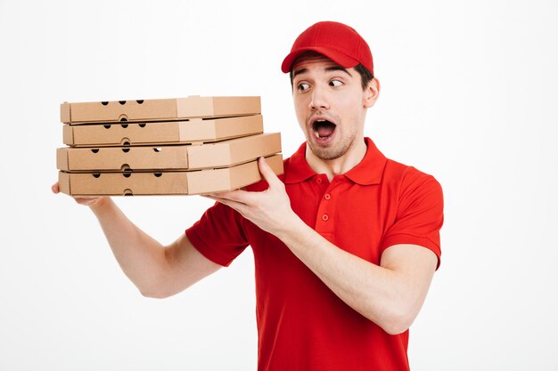 Concessionnaire optimiste en t-shirt rouge et casquette travaillant dans le service de livraison et tenant une pile de boîtes à pizza, isolé sur un espace blanc