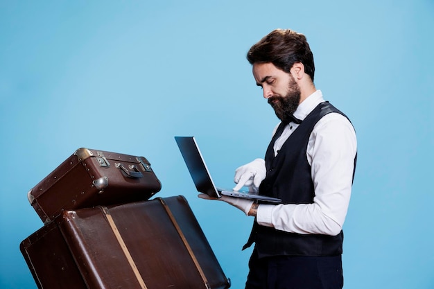 Le concierge de l'hôtel travaille sur un ordinateur portable et se tient à côté d'une pile de bagages en studio, vérifiant la liste des clients ayant réservé en ligne. Un jeune homme travaillant comme chasseur assure une bonne expérience.