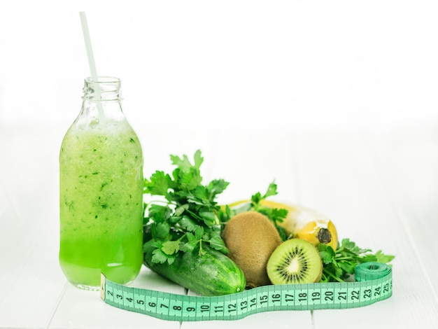 Concombre, kiwi, banane, persil et une bouteille de smoothie sur une table en bois blanche.