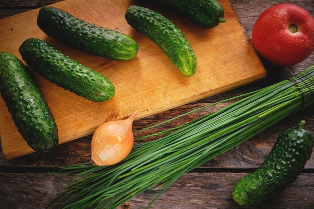 Concombres sur une planche à découper, oignons verts et tomates sur une vieille table en bois.