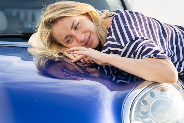 Photo conducteur de femme moyenne embrassant le capot de la voiture après avoir détaillé la publicité d'assurance automobile de polissage