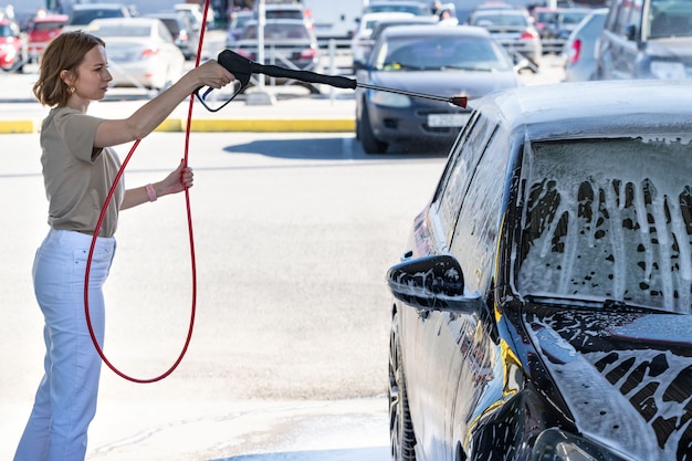 Conducteur de jeune femme se laver au lavage manuel de voiture, nettoyage avec de la mousse, eau sous pression. Lave-auto en libre-service