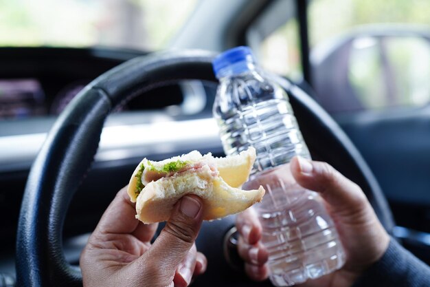 Une conductrice asiatique tient de l'eau froide et du pain sandwich pour manger et boire dans une voiture dangereuse et risque un accident