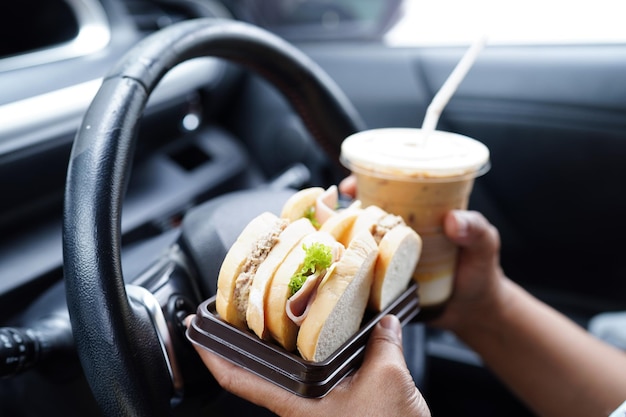 Une conductrice asiatique tient une tasse de café glacé et du pain sandwich pour manger et boire dans la voiture dangereuse et risque un accident