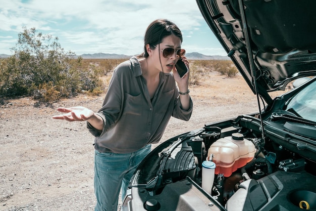 conductrice debout sur la route par une journée ensoleillée à côté de la voiture noire. jeune femme avec des lunettes de soleil utilisant un téléphone portable appelant à l'aide lors de l'accident de voiture en panne sur l'autoroute.