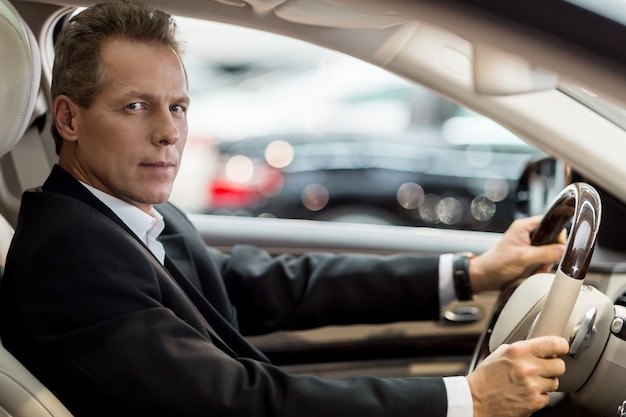 Photo conduite confiante. vue latérale d'un homme âgé confiant en tenue de soirée assis dans une voiture et regardant la caméra