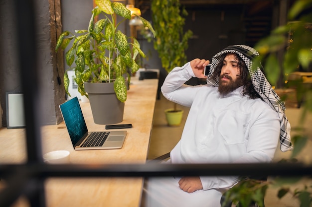 Conférence. Homme d'affaires arabe travaillant au bureau, centre d'affaires utilisant un appareil, gadget. Mode de vie saoudien moderne. L'homme en tenue traditionnelle et écharpe a l'air confiant, occupé, beau. Ethnicité, finances.