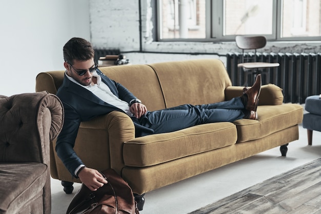 Confiance dans chaque mouvement. Beau jeune homme en costume complet touchant son sac alors qu'il était assis sur le canapé