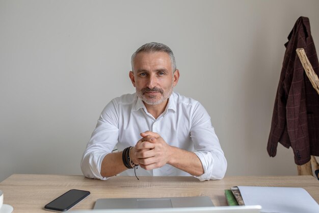 Confiance. Homme d'âge moyen attrayant aux cheveux gris regardant en toute confiance la caméra assis à table dans la chambre contre le mur blanc