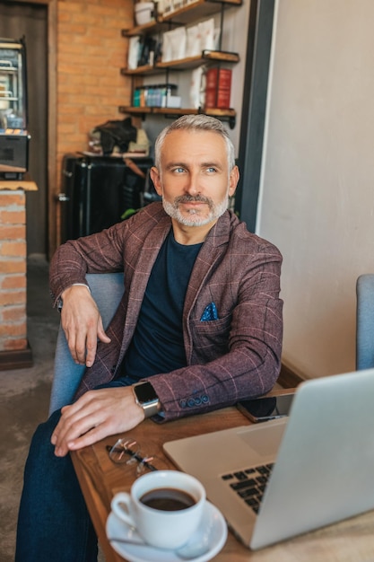 Confiance. Élégant homme aux cheveux gris confiant assis à table avec ordinateur portable et café regardant loin de bonne humeur