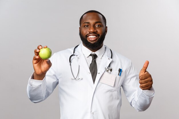 Confiant beau médecin afro-américain recommande de manger des fruits, tenir la pomme verte, sourire heureux et pouce levé, le patient a besoin de vitamines