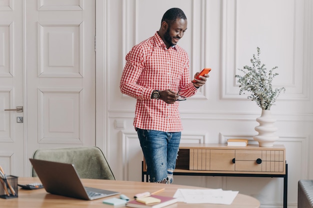 Confiant bel homme d'affaires afro-américain utilisant un mobile debout au bureau pendant la journée de travail