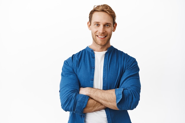 Confiant bel homme rousse avec les bras croisés sur le corps souriant et l'air déterminé comme un professionnel debout dans des vêtements décontractés sur fond blanc