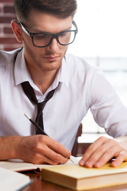 Confiant et créatif. Beau jeune homme en chemise et cravate écrivant quelque chose dans le bloc-notes alors qu'il était assis sur son lieu de travail