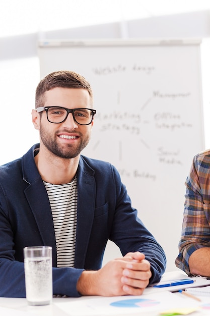 Confiant et créatif. Jeune homme confiant dans des vêtements décontractés intelligents regardant la caméra et souriant alors qu'il était assis sur son lieu de travail avec un tableau blanc en arrière-plan