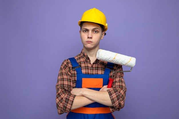 Confiant croisant les mains jeune constructeur masculin portant l'uniforme tenant la brosse à rouleau