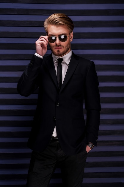 Confiant Dans Son Beau Look. Beau Jeune Homme En Tenue De Soirée Ajustant Ses Lunettes De Soleil Et Tenant Une Main Dans Les Poches En Se Tenant Debout Sur Fond Rayé