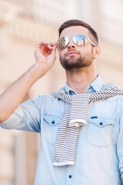 Photo confiant dans son look tendance. beau jeune homme ajustant ses lunettes de soleil tout en se tenant à l'extérieur