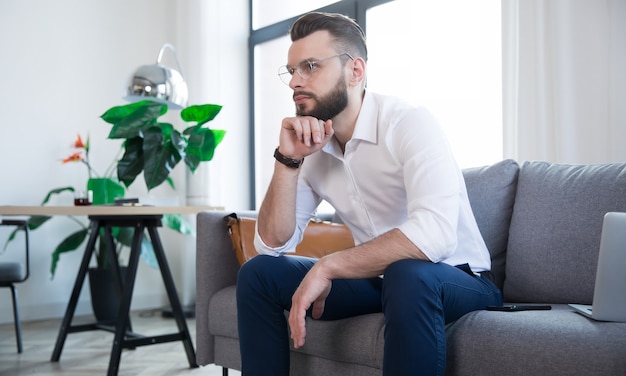 Confiant élégant bel homme d'affaires barbu dans des verres et des vêtements intelligents formels est assis sur le canapé dans le bureau ou la chambre moderne