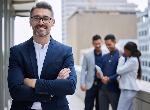 Confiant avec une grande équipe derrière moi Portrait recadré d'un bel homme d'affaires mature debout à l'extérieur avec les bras croisés avec ses collègues en arrière-plan