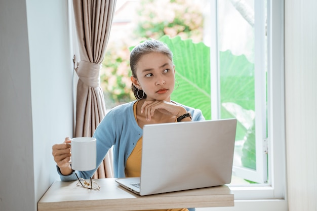 Confiant jeune femme en vêtements décontractés intelligents travaillant sur un ordinateur portable alors qu'il était assis près de la fenêtre à la maison