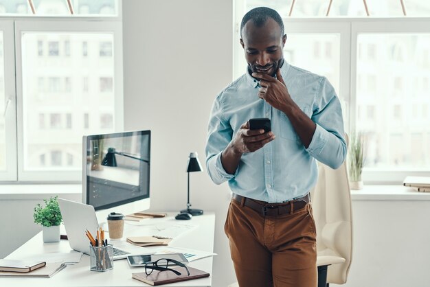 Confiant jeune homme africain en chemise utilisant un téléphone intelligent et souriant tout en travaillant au bureau