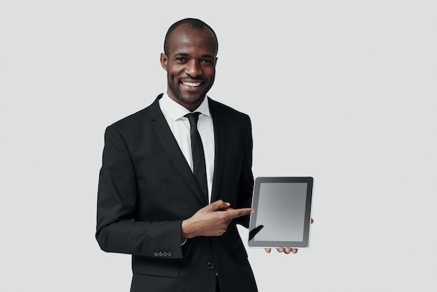 Confiant jeune homme africain en tenue de soirée pointant l'espace de copie sur tablette numérique tout en se tenant contre le mur gris