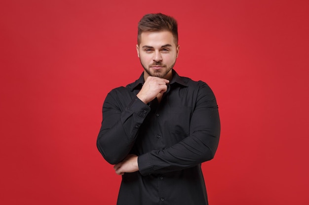 Confiant jeune homme barbu des années 20 en chemise noire classique posant isolé sur fond de mur rouge portrait en studio. Émotions sincères des gens, concept de style de vie. Maquette de l'espace de copie. Mettez la main appuyée sur le menton.