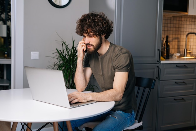 Confiant jeune homme indépendant parlant au téléphone portable assis à table avec un ordinateur portable dans la cuisine avec un intérieur moderne regardant à l'écran