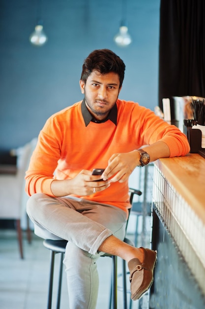 Confiant jeune homme indien en chandail orange assis au comptoir du café avec un téléphone portable à portée de main