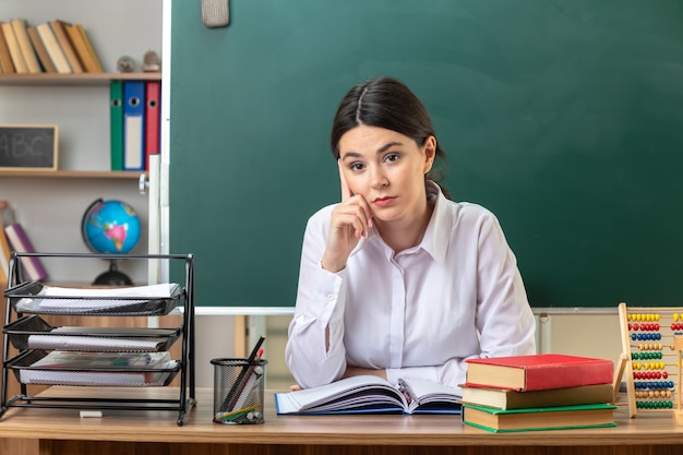 Confiant mettant le doigt sur la joue jeune enseignante assise à table avec des outils scolaires en classe