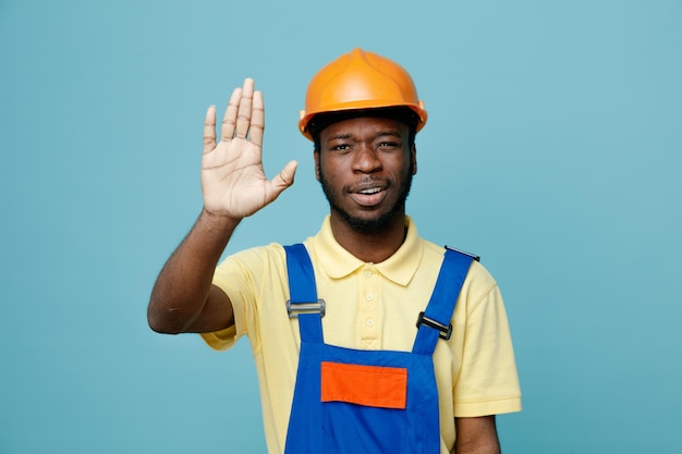 Confiant montrant geste jeune constructeur afro-américain en uniforme isolé sur fond bleu