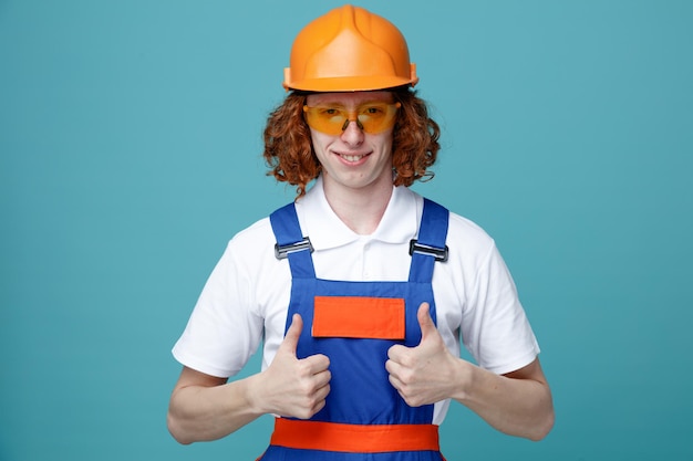 Confiant montrant les pouces vers le haut jeune constructeur homme en uniforme isolé sur fond bleu