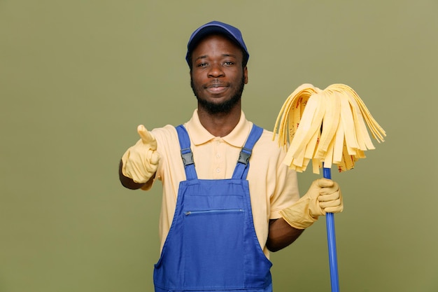 Confiant montrant les pouces vers le haut tenant une vadrouille jeune homme nettoyant afro-américain en uniforme avec des gants isolés sur fond vert