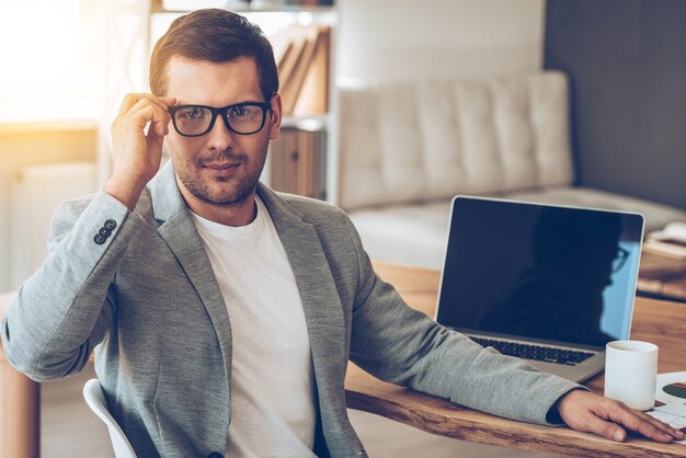 Confiant et réussi. Beau jeune homme ajustant ses lunettes et regardant la caméra alors qu'il était assis sur son lieu de travail