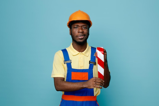 Confiant tenant du ruban adhésif jeune constructeur afro-américain en uniforme isolé sur fond bleu