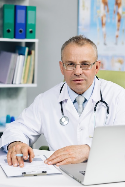 Confiant vieux médecin-chef mâle mature en uniforme médical blanc dans des verres assis sur le lieu de travail.