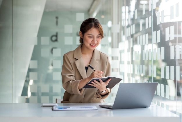 Confiante et belle jeune femme d'affaires asiatique assise en prenant des notes dans le bloc-notes avec un ordinateur portable au bureau