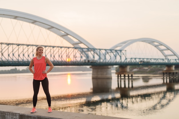 Confiante forte fille blonde sportive mignonne debout sur la jetée près de la rivière après l'entraînement tôt le matin.