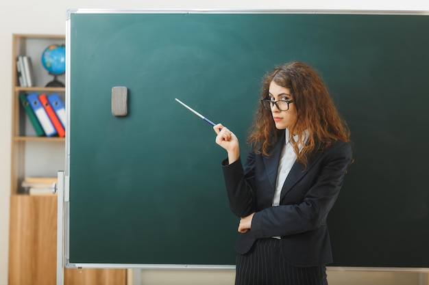 confiante jeune enseignante debout devant des points de tableau noir au tableau noir avec un pointeur dans la salle de classe