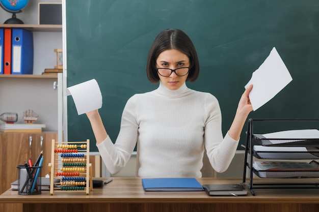 confiante jeune enseignante portant des lunettes déchirer du papier assis au bureau avec des outils scolaires en classe