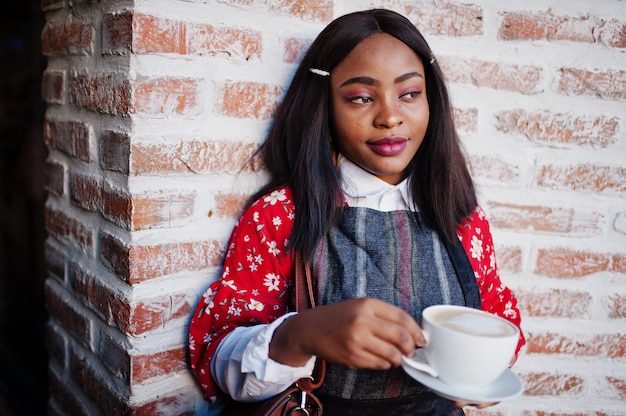 Ð¡ confiante jeune femme afro-américaine dans des vêtements décontractés intelligents au café avec une tasse de boisson chaude à la main.