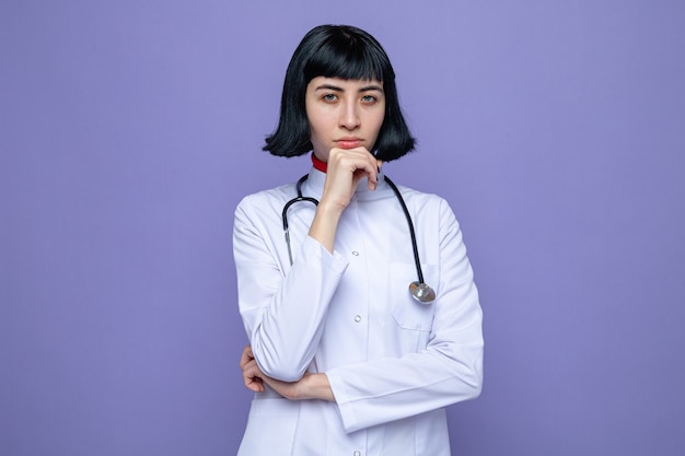 Confiante jeune jolie fille caucasienne en uniforme de médecin avec stéthoscope mettant la main sur son menton et regardant à l'avant