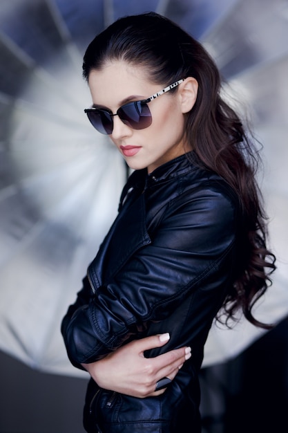 Confiante et sexy jeune femme brune posant en veste en cuir noir et lunettes de soleil