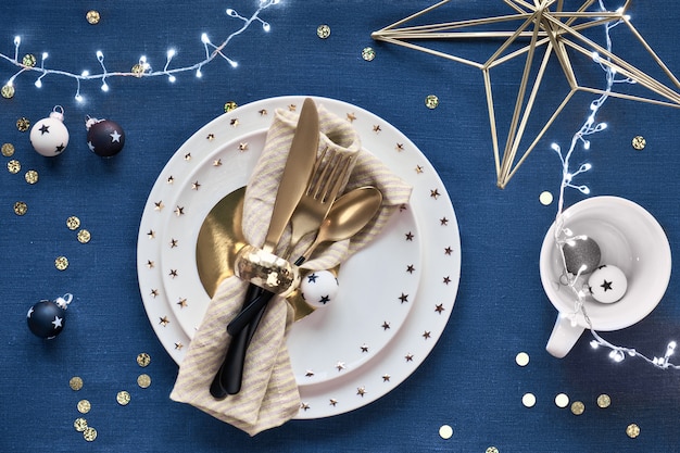 Configuration de table de Noël avec assiette blanche et ustensiles dorés et décorations dorées. Mise à plat, vue de dessus sur bleu classique. Fond textile lin bleu foncé.