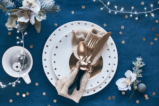 Configuration de table de Noël avec assiette blanche et ustensiles dorés et décorations dorées. Mise à plat, vue de dessus sur fond de textile en lin bleu classique.