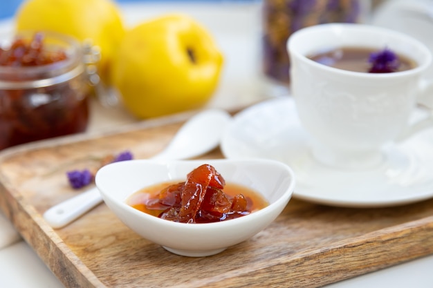 Confiture de coings maison dans un bol en porcelaine avec une tasse de thé sur un plateau en bois.